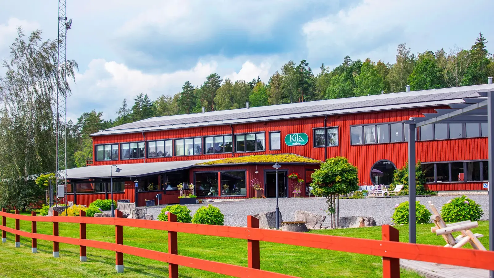 Kila Möblers butik i Kiladalen. En stor faluröd byggnad med parkering på framsidan omgiven av grön natur.